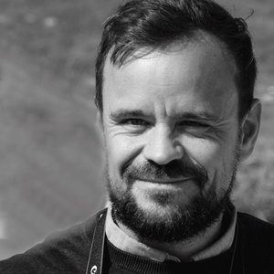 Mann med skjegg og smil, iført svart genser og kamera rundt halsen, står ute i naturlig lys og det er bra.