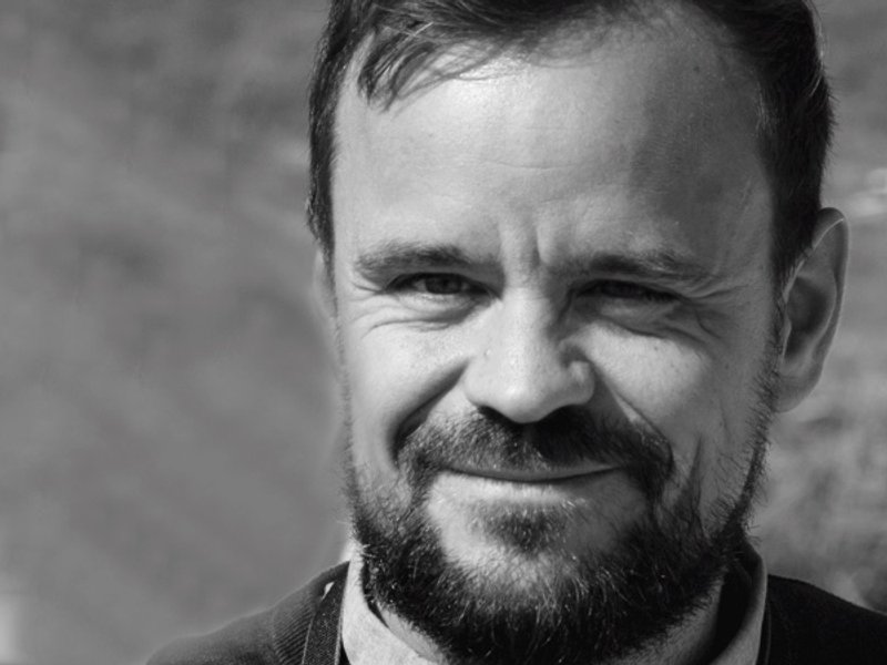 Mann med skjegg og smil, iført svart genser og kamera rundt halsen, står ute i naturlig lys og det er bra.